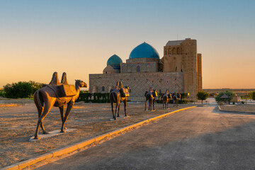 Turkestan, Kazakhstan - June 03, 2025: Scenic early morning sunrise view of the historical Mausoleum of Khoja Ahmed Yasawi (Қожа Ахмет Ясауи) with a bronze camel statue in the foreground © Bilal