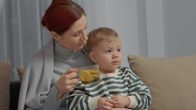A sick woman holds her little boy while they sit together in a cozy living room. She drinks tea as part of her treatment for cold symptoms and recovery.