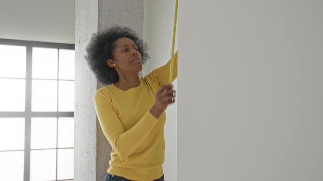 Woman in yellow sweater measures a white wall holding and pulling a tape measure against the surface in studio; concentration planning preparation.