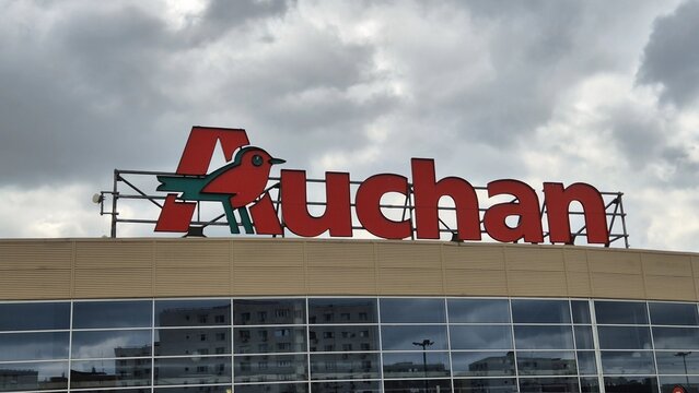 The large, red "Auchan" logo with its signature green and red bird sits atop a modern building under a cloudy, overcast sky. April 10, 2026 in Bucharest, Romania.