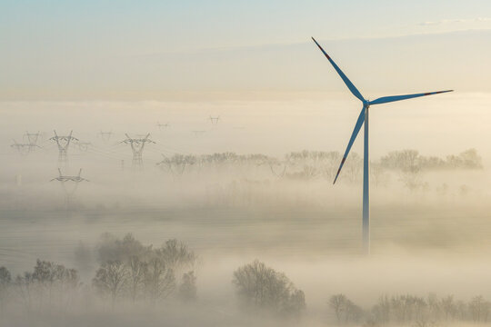 Aerial view of a solitary wind turbine piercing through a dense fog blanket, with distant power lines adding to the ethereal landscape, Wislina, Pomeranian Voivodeship, Poland.