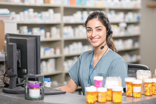 A smiling pharmacist wearing scrubs and a headset works at a computer in a pharmacy surrounded by prescription medicine bottles.
