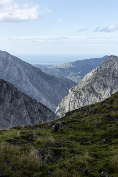 View of rugged, sun-kissed mountain peaks rise majestically from verdant slopes under a soft sky, a serene panorama of nature's grandeur, Picos de Europa, Asturias, Spain.