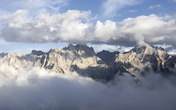 View of jagged peaks pierce through a sea of swirling clouds, sunlight catching the stark rock faces, creating a dramatic contrast of light and shadow, Picos de Europa, Asturias, Spain.