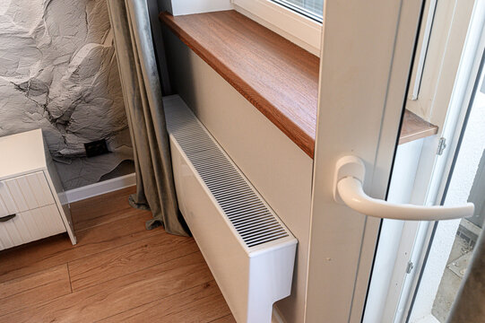 A white convector heater under a wooden window sill, beside a glass door with a white handle; light wood flooring, textured grey wall, white bedside table, beige curtain partially visible.