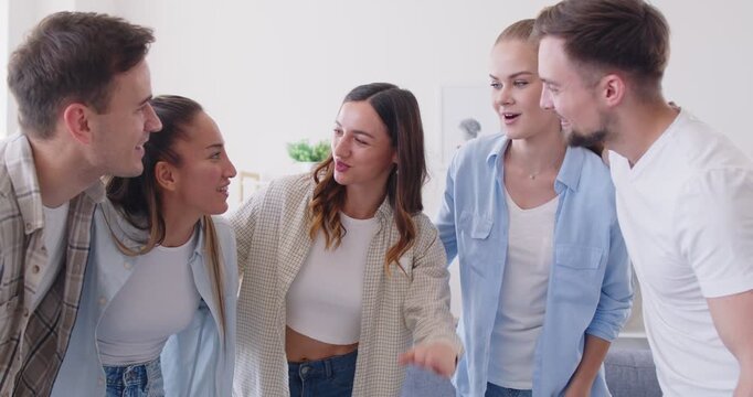 Young smiling positive woman sharing news, telling funny story or joke to her friends who listening and laughing. Group of happy people standing in circle, hugging, showing closeness and support.