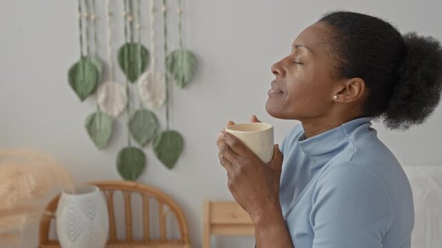 Woman holds coffee cup to chest with closed eyes on bed by rattan headboard and leaf wall hanging in studio; morning serenity.