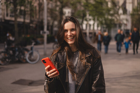 Cheerful young woman in leather jacket laughing while using red smartphone on busy city street, happy emotional portrait in urban environment, digital communication and lifestyle concept.