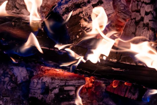 Burning wood in a fire pit, night setting. Flames in yellow, orange, and white flicker across charred logs. Glowing embers and ash visible beneath and around the wood. Dark, charred surfaces.