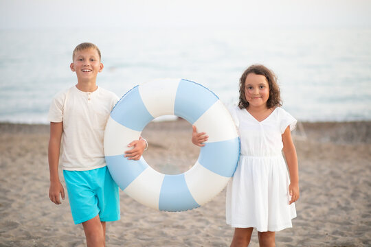 Two children playfully peeking through a blue and white inflatable ring on a sandy beach, wearing light summer clothes, with a blurred natural background.
