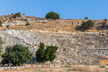 Naklejka premium Ruined Roman Theatre of Patara ancient city. A historic stone amphitheater ruins with terraced seating, an arched entrance, and weathered walls sits in a sunlit landscape. Turkey