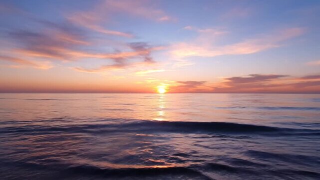 Soft pastel sunrise over calm ocean waters with wispy clouds and gentle waves washing onto the shore. The sun reflects on the sea surface with pink and orange sky colors at dawn.