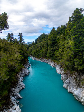View of of a vibrant turquoise river slicing through a dense, emerald forest, the rugged banks a stark contrast under a cloudy sky, Hokitika Gorge, New Zealand.
