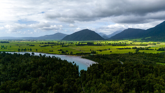 Aerial view of a turquoise river snaking through a verdant valley, flanked by dark forests and majestic mountains beneath a cloudy sky, Hokitika Gorge, West Coast Region, New Zealand.