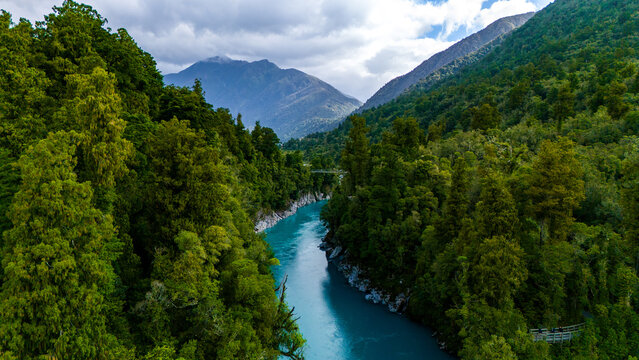Aerial view of the Hokitika Gorge's turquoise waters carve a path through the dense, vibrant green forest under a sky of soft, diffused light, Hokitika Gorge, West Coast Region, New Zealand.