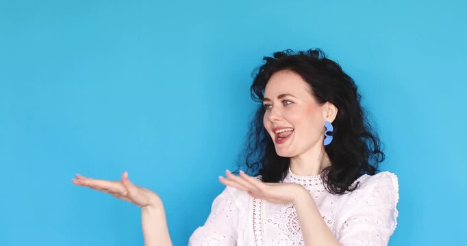 Cheerful curly haired woman dancing gracefully in studio against blue background, happy girl in white lace blouse lip-syncing and moving with joy, positive emotions and musical vibes.