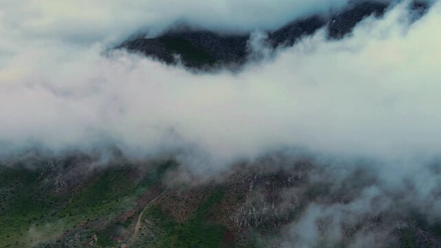 Misty clouds drift over rugged mountain peaks. Sunlight glows soft against the misty veil. Vegetation clings to steep, mossy slopes. Mist swells and recedes, revealing hidden peaks