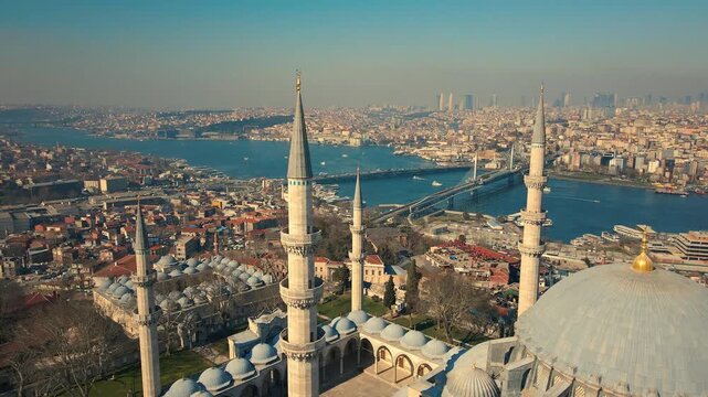 Blue dome gleams under golden sky. Minarets rise above ancient cityscape. Sunlight bathes mosque in serene glow. Crowds below move gently through streets. City sprawls behind, hazy with distance