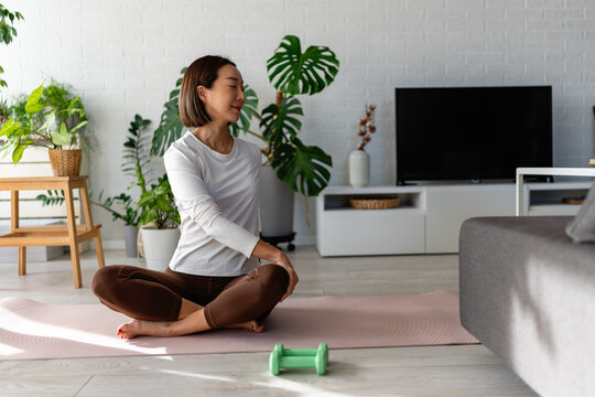 East Asian woman sitting cross-legged on a yoga mat, gently twisting her torso during a home workout. Calm, mindful moment with indoor plants and soft daylight, promoting wellness and balance