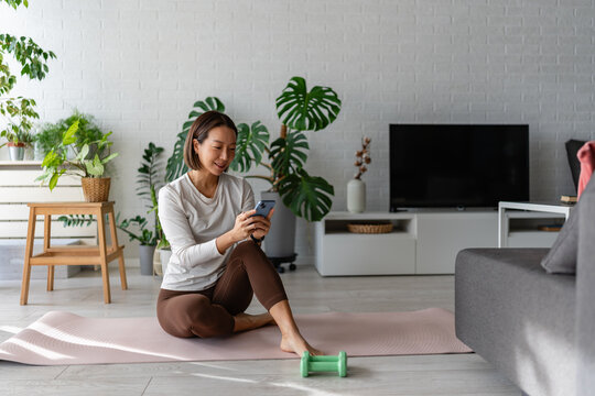 East Asian woman sitting on a yoga mat at home, using her smartphone after a workout. Calm, focused moment with indoor plants and soft daylight, reflecting a balanced, modern wellness lifestyle