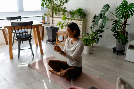 East Asian woman sitting on a yoga mat at home, using her smartphone after a workout. Calm, focused moment with indoor plants and soft daylight, reflecting a balanced, modern wellness lifestyle