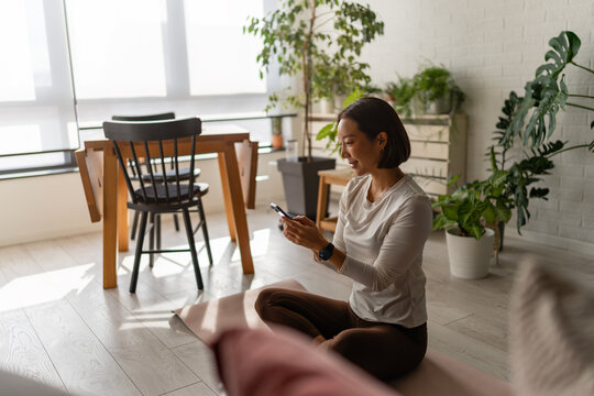 East Asian woman sitting on a yoga mat at home, using her smartphone after a workout. Calm, focused moment with indoor plants and soft daylight, reflecting a balanced, modern wellness lifestyle
