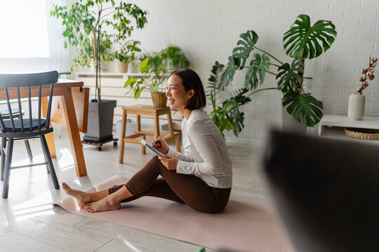 East Asian woman sitting on a yoga mat at home, writing notes in a notebook after a workout. Calm, focused wellness moment with indoor plants and natural light in a cozy living space