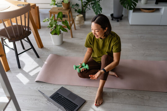 Asian woman fitness instructor exercising on a yoga mat with dumbbells while streaming a workout on laptop. Online coaching scene showing communication, motivation and active lifestyle.