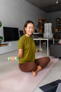 Asian woman exercising at home on a yoga mat with dumbbells while following a laptop workout. Healthy lifestyle scene showing motivation, focus and active living indoors.