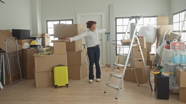 Woman with arms outstretched among stacked moving boxes, yellow suitcase and ladder in a building; fresh start joy.