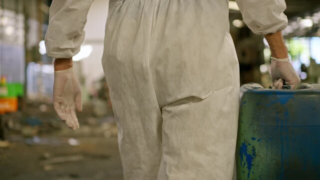 Hazardous waste industrial worker protective suit with safety gloves handling chemical barrel and plastic drum chemical container inside factory interior for waste management, industrial safety