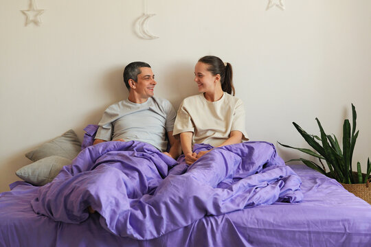 Two people are sitting on a bed covered with purple blankets. They are smiling and looking at each other in a bright room with plants