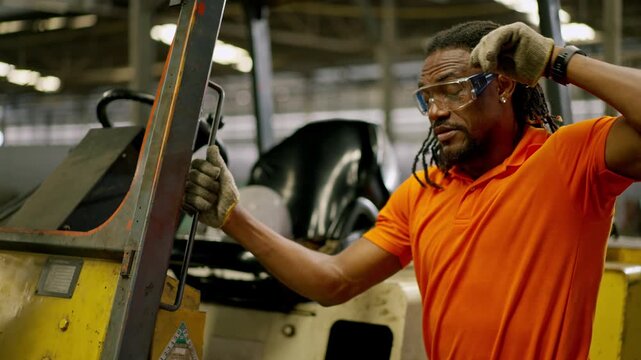 Factory worker portrait in industrial warehouse beside forklift wearing safety goggles and glove, orange shirt maintenance labor, tired worker wiping sweat during manual work near forklift
