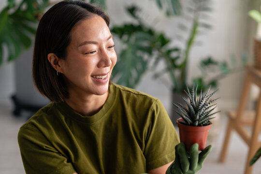 Young East Asian woman smiling while holding a small potted succulent. Cozy indoor gardening moment with natural light, plant care, and relaxed home lifestyle