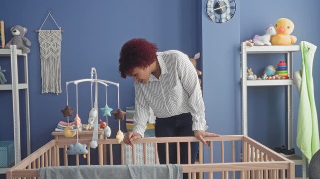 Woman leans over crib, hand on rail in studio nursery smiling at baby mobile softly; motherhood tenderness.