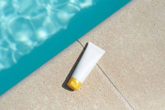 Sunblock or sunscreen in cosmetic tube sits on the edge of a swimming pool. The clear blue water reflects sunlight. Summer vacation and skin care, protect cosmetics concept. Skin health