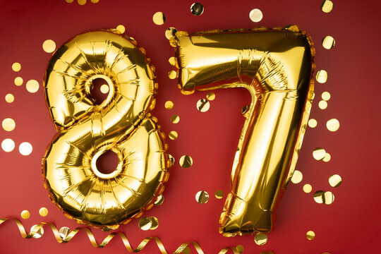 87th birthday celebration. Gold festive balloons number 87 year anniversary on red background with confetti. Birthday or wedding event.