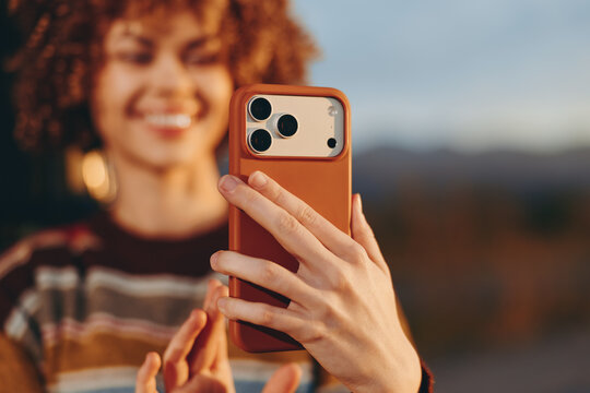 Woman lifestyle selfie outdoors, smiling with curly hair blurred behind, holding smartphone with orange case, close foreground hands, warm golden light, relaxed casual mood, evening breeze