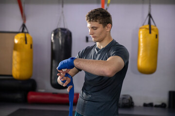 Adult male wearing grey tee, shorts wrapping blue hand wraps at boxing gym with heavy bags © wavebreak3