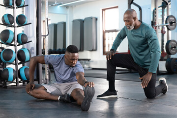 Senior male trainer kneeling beside male client stretching on mats by ball rack, both wearing shoes © wavebreak3