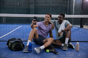 Diverse male friends sitting on blue court, holding smartphone and yellow ball, posing near racket © wavebreak3