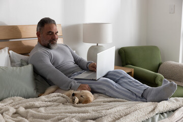 Mature adult man reclining on bed in striped pajamas, using silver laptop and petting golden puppy © wavebreak3
