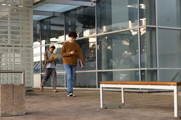 Two male students walking beside glass building on campus checking smartphone holding folders © wavebreak3