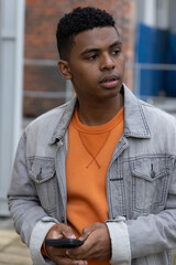 African American teen holding smartphone and looking right by brick wall, wearing denim jacket © wavebreak3