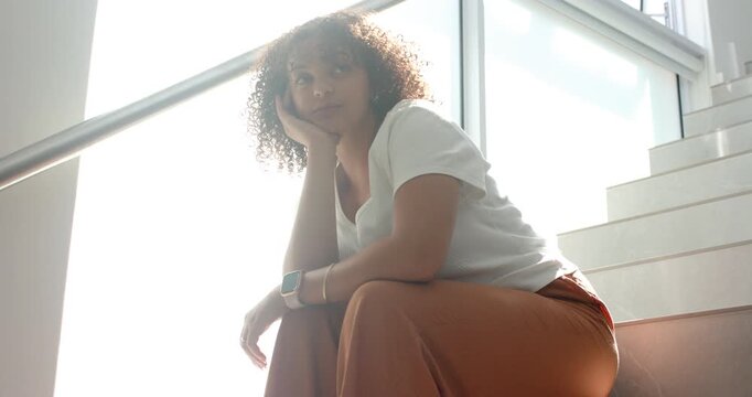 African American woman sitting on stairs beside backlit window and rail, holding chin, reflecting