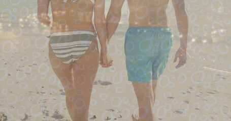 Fototapeta premium Walking couple holding hands on beach with striped bikini bottoms, aqua trunks, footprints, overlay