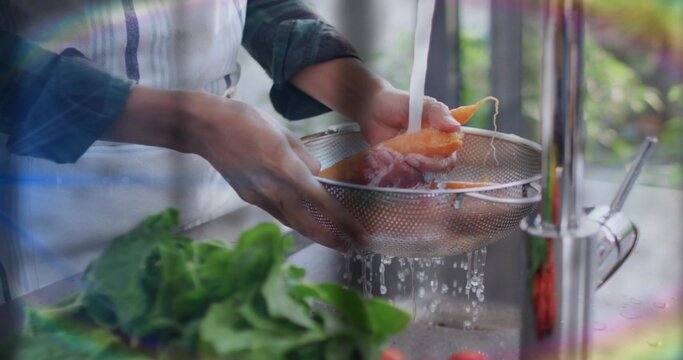 Rinsing cook's hands holding mesh colander with orange pieces under sink tap, in apron, plaid-shirt