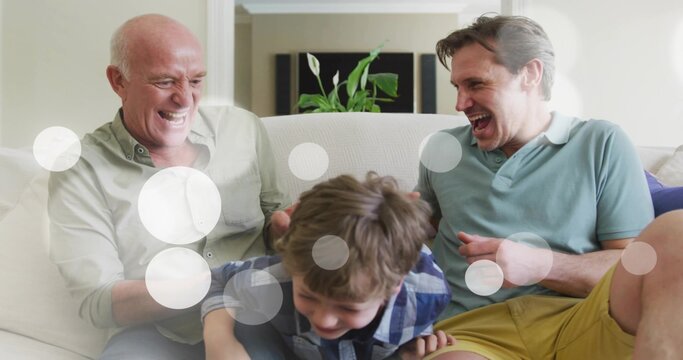 Male three-generation family laughing as granddad and father reaching toward child on sofa for play