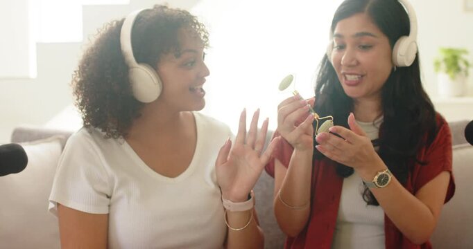 Female duo, right holding jade roller, demonstrating on couch wearing headphones, mic recording
