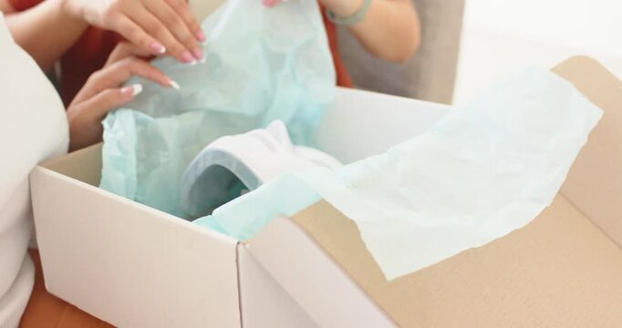 Diverse female friends receiving box on couch, unwrapping tissue and revealing mask to inspect